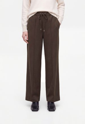 Persoon draagt donkerbruine wijde broek met trekkoord in de taille, lichtbeige trui en zwarte schoenen met vierkante neus, staat met handen in de zakken.