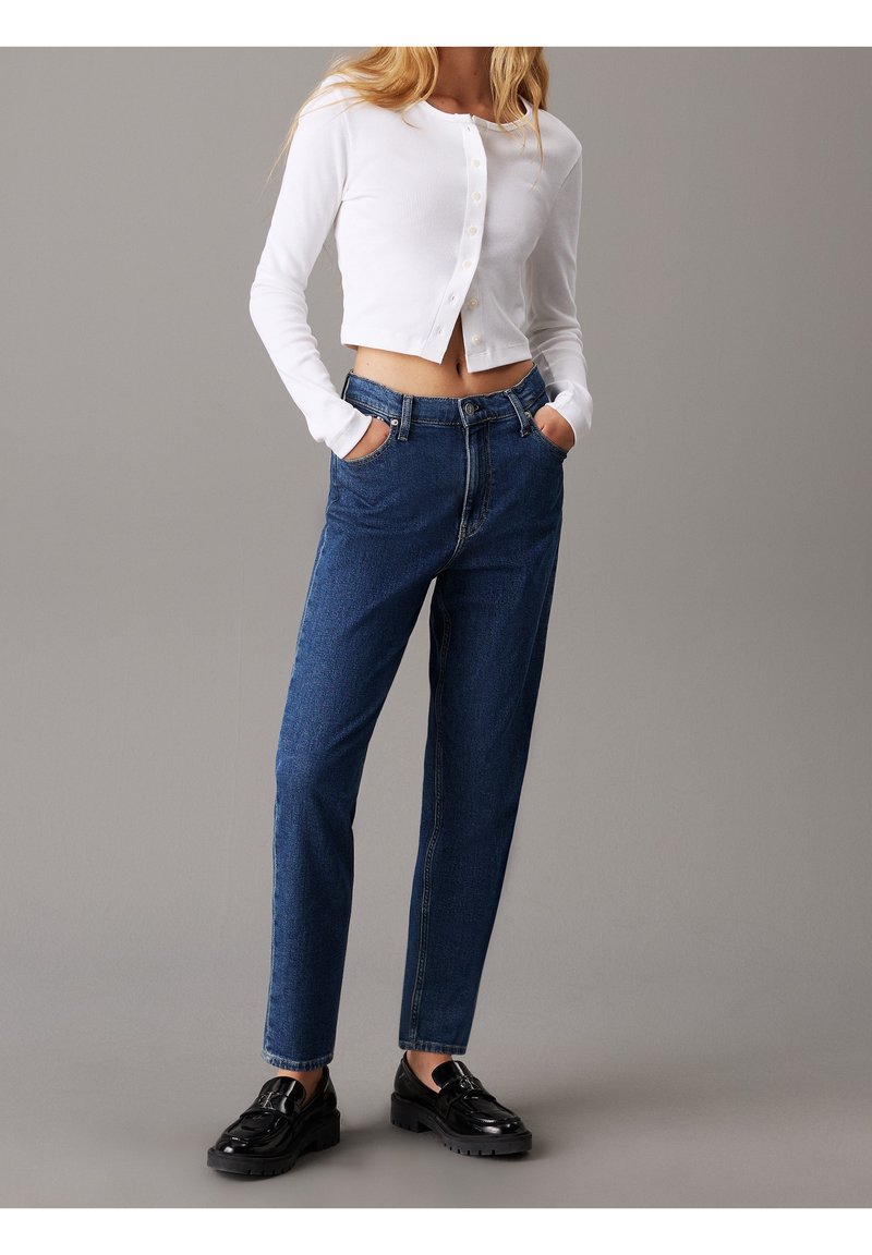 White ribbed long-sleeve cropped cardigan with button closure, paired with high-waisted dark blue denim jeans and black platform shoes.