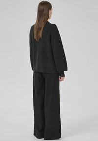 Eine Frau mit langen braunen Haaren trägt einen dunkelgrauen Strickpullover und weit geschnittene schwarze Hosen, während sie mit dem Gesicht von einer schlichten grauen Hintergrund abgewandt steht.
