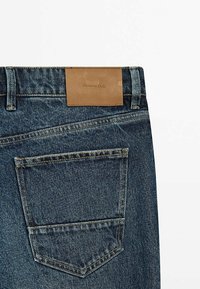 Hintere Tasche einer blauen Jeans aus Denim mit braun genähtem Leder-Etikett der Marke "Massimo Dutti" am Bund vor weißem Hintergrund.