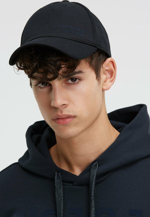 Young man with curly hair wearing a dark baseball cap and matching hoodie, looking directly at the camera with a neutral expression.