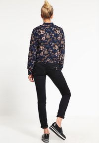 Blommönstrad marinblå bomberjacka med långa ärmar och svarta slim-fit jeans. Svarta slip-on skor med vita sulor. Slät tygtextur.