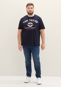 Granatowa bawełniana koszulka z krótkim rękawem z białym i czerwonym napisem; zestawiona z niebieskimi dżinsami i białymi trampkami.