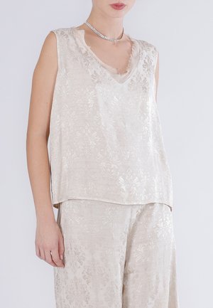 Donna che indossa un top senza maniche color crema in broccato e pantaloni a gamba larga abbinati con motivo damascato floreale e una collana choker d'argento.