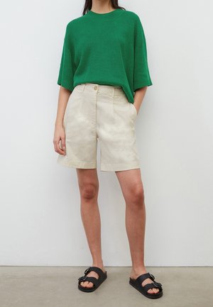 Grüner, strukturierter Strickpullover mit kurzen Ärmeln, kombiniert mit beigen, taillierten Shorts. Schwarze Sandalen mit einer dekorativen Schnalle vervollständigen das Outfit.