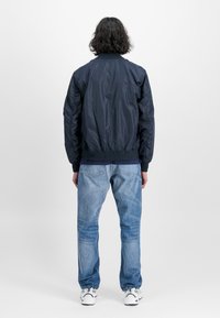 Marinebombejakke med en jevn tekstur, ribbestrikkede mansjetter og nederkant, og en tettsittende design, sammen med lyseblå jeans og hvite joggesko.