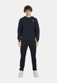 Felpa blu navy con polsini a coste, scollatura rotonda e piccola etichetta arancione sul petto. Abbinata a pantaloni blu navy e sneakers bianche.