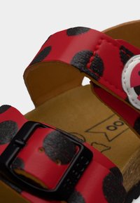 Κόκκινο σανδάλι με μαύρα πουά, δύο ρυθμιζόμενα λουράκια που στερεώνονται με μαύρη αγκράφα και φυσική υφασμάτινη εσωτερική σόλα. Ελαφρύ σχέδιο.