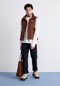 Bruine, geïsoleerde vest met een hoge kraag; gedragen over een witte blouse met lange mouwen. Zwarte broek, beige wandelschoenen en een bruine schoudertas.