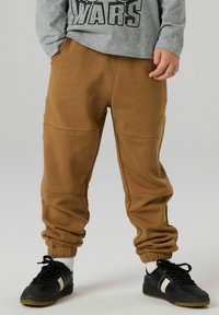Brązowe spodnie dresowe wykonane z bawełnianej mieszanki, z elastycznymi mankietami, szwami po bokach i luźnym krojem. Noszone z czarnymi tenisówkami i białymi skarpetkami.