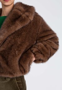 Giacca in ecopelliccia marrone con collo ampio, maniche oversize e texture morbida. Il modello indossa un maglione verde e pantaloni oliva.