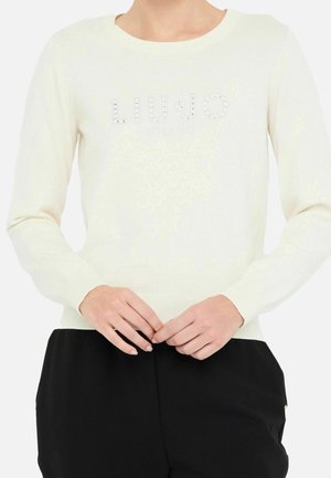 Cream long-sleeve sweater with "LIU JO MILANO" logo in small crystals worn with black pants, hands gently clasped in front.