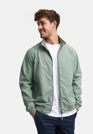 Uśmiechnięty mężczyzna o krótkich, kręconych włosach, ubrany w jasnozieloną bluzę na zamek, białą koszulkę i ciemnoniebieskie spodnie, z rękami w kieszeniach, patrzący w lewo.