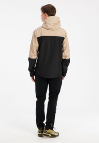 Vesito vedenkestävä takki mustassa ja beigeväri, jossa on huppu, säädettävät hihansuut ja sileä pinta. Yhdistetty mustiin housuihin ja vaelluskengiin.