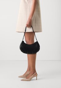 Black leather handbag with curved design, adorned with silver studs on the sides. Short handle, paired with beige high-heeled shoes.
