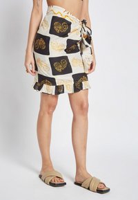 Gonna avvolgente in crema con motivo ostrica nero e giallo, presenta un orlo arricciato e un cinturino annodato in vita. Indossata con sandali piatti color cammello.