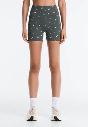 Nő sötétzöld, magas derekú kerékpáros rövidnadrágot visel apró virágmintával, fehér zoknival és törtfehér tornacipővel, fehér háttér előtt áll.