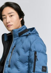 Blaue, gepolsterte, isolierte Jacke mit einem Reißverschluss vorne, hohem Kragen und Kapuze. Verfügt über eine Seitentasche mit Reißverschluss und schwarzen Akzenten.