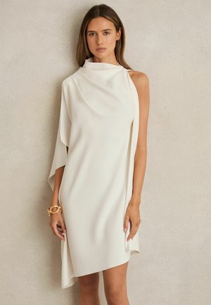 Vrouw draagt een mouwloze asymmetrische witte jurk met één gedrapeerde mouw en een gouden armband, staand tegen een beige muur.