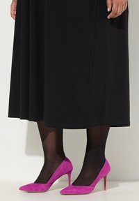 Des chaussures à talons hauts en suede rose avec un design à bout pointu, portées avec des collants opaques noirs, associées à une longue jupe noire.