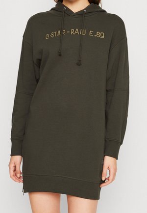 Kvinde iført en olivengrøn oversized hoodie-kjole med "G-STAR RAW E.89" broderet på brystet og snoretræk i hætten.