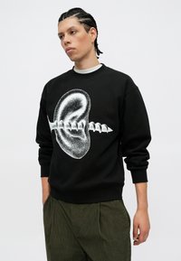 Μαύρη ζακέτα με μεγάλη λευκή γραφιστική απεικόνιση ενός αυτιού που είναι μπλεγμένο με μια βίδα. Διαθέτει ribbed μανσέτες και στρογγυλή λαιμόκοψη.