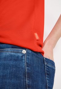 Personne portant un jean bleu en denim, une main dans la poche, et une chemise rouge avec une petite étiquette en tissu sur la couture latérale.