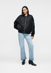 Veste bomber noire avec fermeture éclair, poignets côtelés et poches latérales zippées, portée avec un jean évasé bleu clair et des bottines noires.