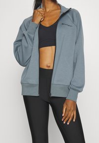 Grå zip-up hoodie i mjukt tyg, med hög krage, ribbade ärmar och en liten logotyp på vänster bröst. Matchas med svarta leggings.