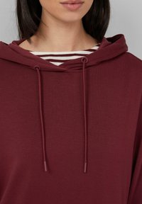 Bordeaux hoodie met een trekkoord, zachte textuur en een ronde hals, gelaagd over een gestreept wit en bruin shirt.