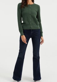 Grön strukturerad kofta med rund halsringning, knappstängning och ribbade ärmar, kombinerad med mörka utsvängda jeans i en helkroppsbild.