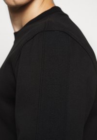 Sudadera negra de manga larga con textura acanalada, cuello redondo y detalle de logotipo en relieve a lo largo del hombro.