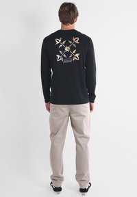 Chemise noire à manches longues avec des motifs floraux et feuilles multicolores dans le dos, associée à un pantalon beige et des baskets noires.