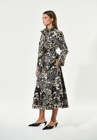 Obleka s cvetličnim vzorcem v črni in bež barvi, z visokim ovratnikom, zategnjenim pasom in žepi, v kombinaciji s črnimi čevlji s koničastimi prsti.