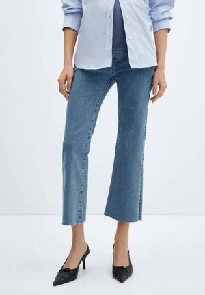 Kobieta ubrana w jasnoniebieskie krótkie dzwony, jasnoniebieską zapinaną na guziki koszulę oraz czarne czubiaste buty na wysokim obcasie, stoi na białym tle.