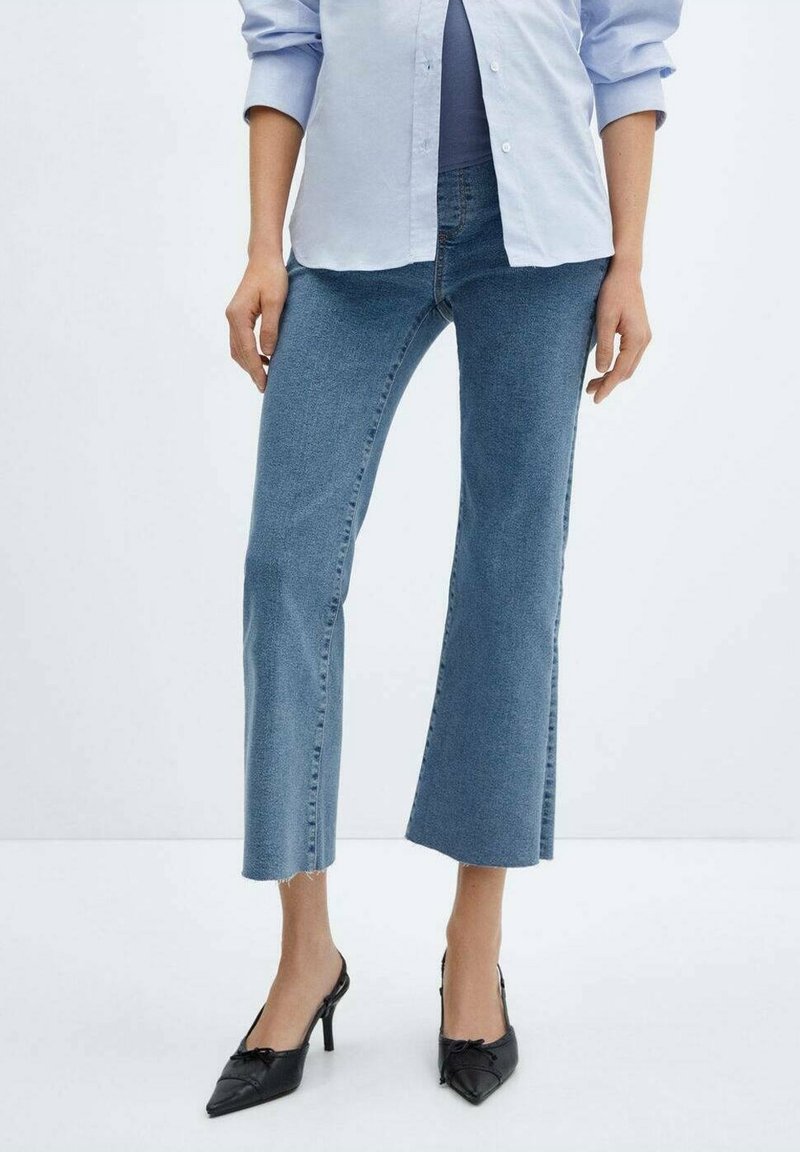 Femme portant un jean flare court bleu clair, une chemise bleu clair boutonnée, et des chaussures noires à talons aiguilles pointues, se tenant devant un fond blanc.