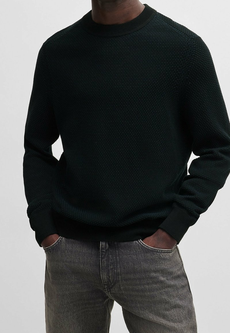 Homme portant un pull noir texturé à manches longues, les mains dans les poches d’un jean gris délavé, sur un fond uni.
