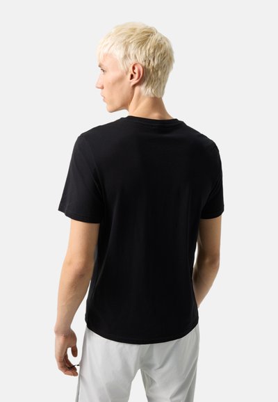 Ung person med kort blondt hår, iført en ensfarvet sort kortærmet t-shirt og lyse bukser, stående med ryggen til.