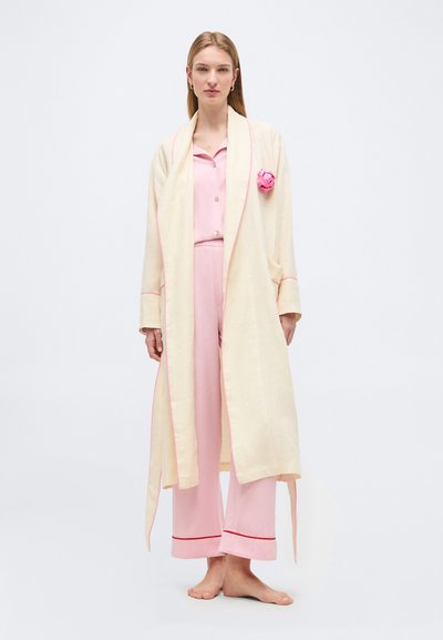 Γυναίκα που στέκεται ξυπόλητη φορώντας ανοιχτό ροζ πιτζάμες και ένα μακρύ κρεμ ρόμπα με ροζ διακοσμητικές λεπτομέρειες και ροζ λουλούδι στην τσέπη στο στήθος.