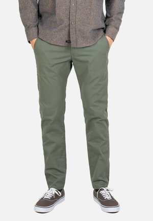 Mann trägt olivgrüne Hose, graues langärmliges Hemd mit Knöpfen und graue Sneakers mit weißen Schnürsenkeln, Hände in den Taschen.