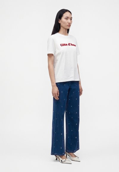Femme aux longs cheveux foncés porte un t-shirt blanc avec un texte rouge, un jean bleu large avec de petits motifs blancs, et des chaussures blanches à talons.