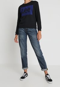Felpa nera con logo "Lee" blu, abbinata a jeans blu medio con risvolto e sneakers nere con dettagli bianchi. Look casual.