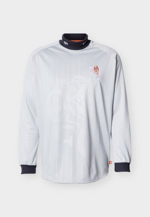 Λευκή αθλητική φανέλα με μακριά μανίκια, μαύρο γιακά και μανσέτες, με έμβλημα κόκκινου λέοντα και αρχικά "CFC" στο στήθος.