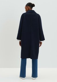 Marineblauer Wollmantel mit lässiger Silhouette, weiten Ärmeln und Kragen, getragen über blauen Schlagjeans. Schlichtes Design, keine bemerkenswerten Akzente.