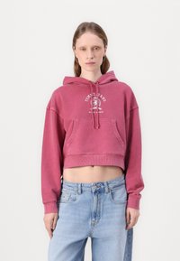 Felpa rosa tagliata, con tasca frontale e cappuccio con cordino. Presenta un logo bianco sul petto. Abbinata a jeans blu chiaro.