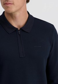 Mann trägt ein dunkelblaues Zip-Polohemd mit geripptem Muster und einem kleinen "Pierre Cardin"-Logo auf der linken Brust.