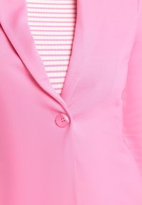 Blazer rosa con una texture liscia, caratterizzato da un colletto a punta e un dettaglio centrale con bottone, indossato sopra una maglietta a righe rosa e bianche.