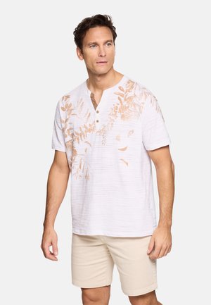 Hombre que lleva una camiseta henley de manga corta blanca con estampado de hojas beige y pantalones cortos beige claro, de pie y mirando ligeramente hacia un lado.