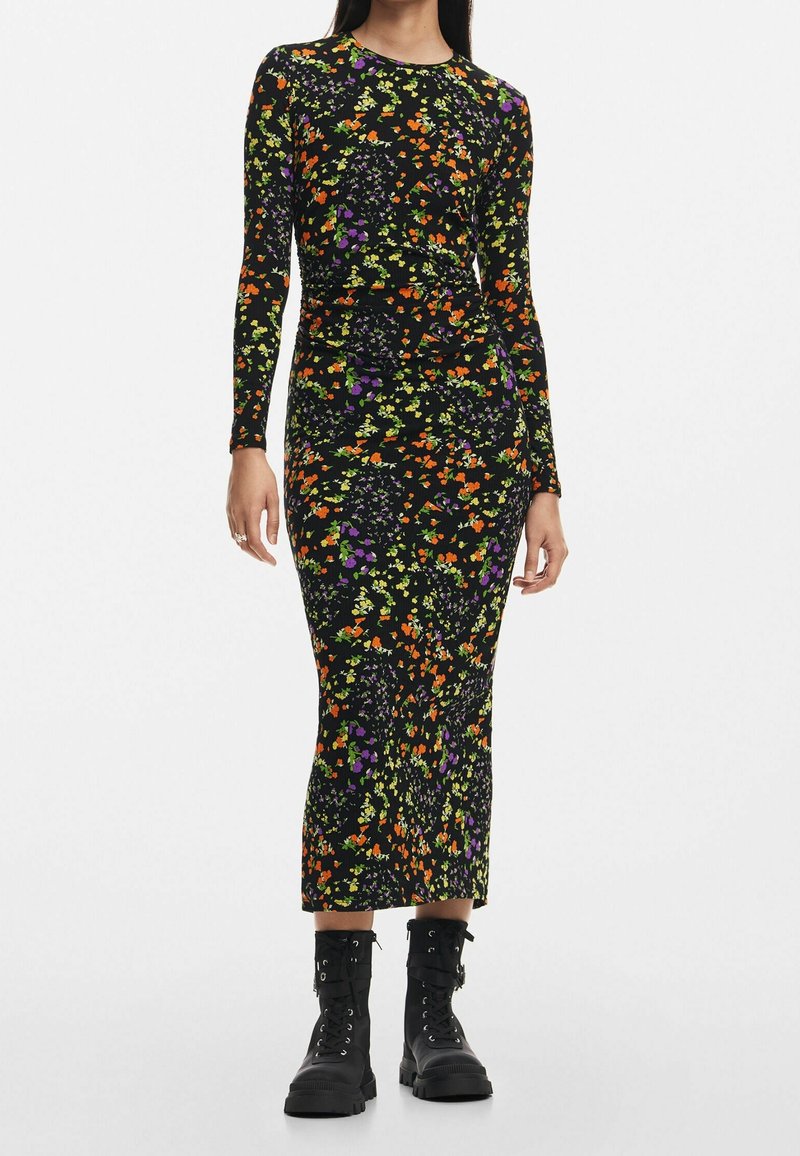 Femme portant une robe noire cintrée à manches longues avec un imprimé floral orange, jaune et violet, associée à des bottes de combat noires.