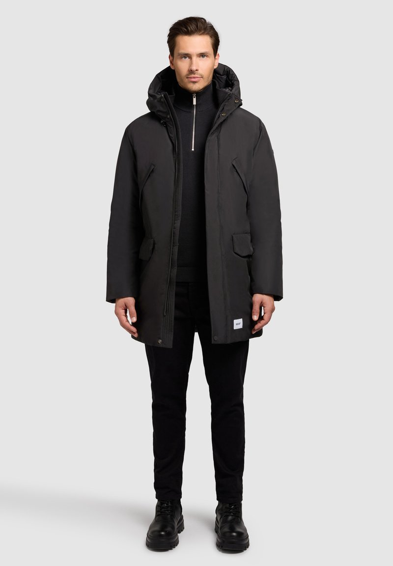 Sort parka-jakke med hætte, med lynlås foran, to forlommer og et struktureret stof. Båret over en sort sweater og bukser.